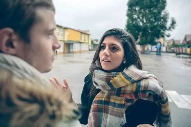 A woman in a plaid scarf and coat looks distressed while talking to a man outdoors on a rainy day.
