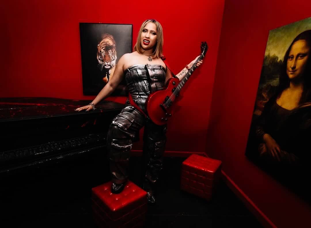 A woman with a guitar in a vibrant red-walled art studio, surrounded by iconic artwork including the Mona Lisa.