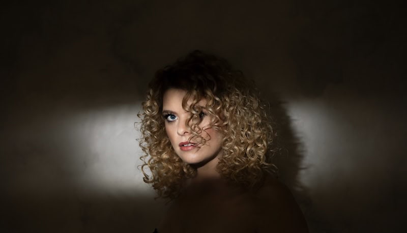Close-up of confident woman with curly blonde hair and piercing gaze, dramatic lighting, portrait style.