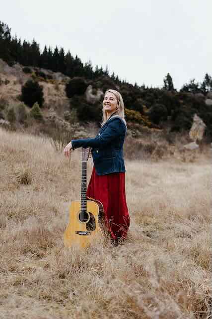 Keily Smith 1 A woman in a red dress and denim jacket standing in an open field with an acoustic guitar, surrounded by hills and trees.