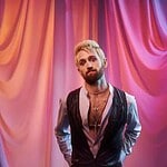 Male artist with a beard and styled hair standing in front of colorful draped curtains.