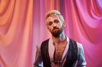Male artist with a beard and styled hair standing in front of colorful draped curtains.