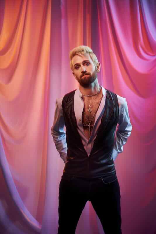 Male artist with a beard and styled hair standing in front of colorful draped curtains.