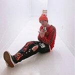 Young man sitting on the floor in a white hallway, wearing a red beanie and colorful streetwear.