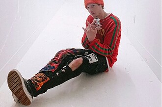 Young man sitting on the floor in a white hallway, wearing a red beanie and colorful streetwear.