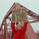 Woman holding a sign asking "What is the meaning of life?" under a bridge.