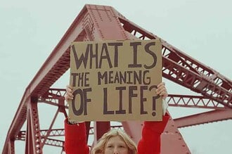 Woman holding a sign asking "What is the meaning of life?" under a bridge.