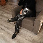 Alxandr sitting thoughtfully on a sofa, wearing stylish black leather pants and boots, in a cozy ind.