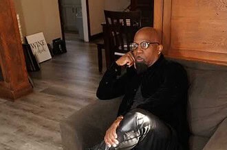 Alxandr sitting thoughtfully on a sofa, wearing stylish black leather pants and boots, in a cozy ind.