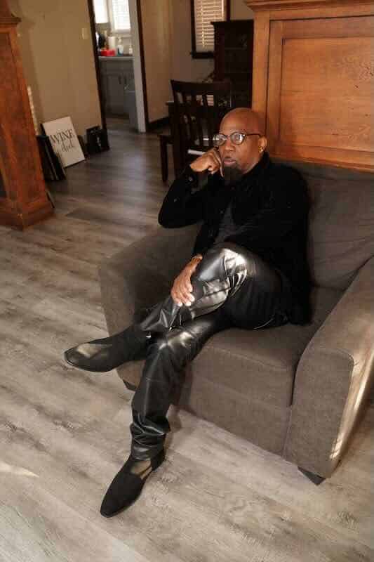 Alxandr sitting thoughtfully on a sofa, wearing stylish black leather pants and boots, in a cozy ind.