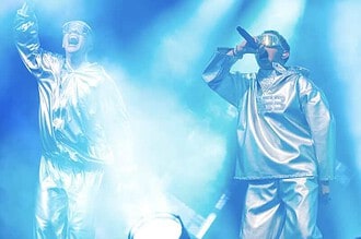 VÆB and GamerBoi performing on stage with futuristic outfits and microphones.